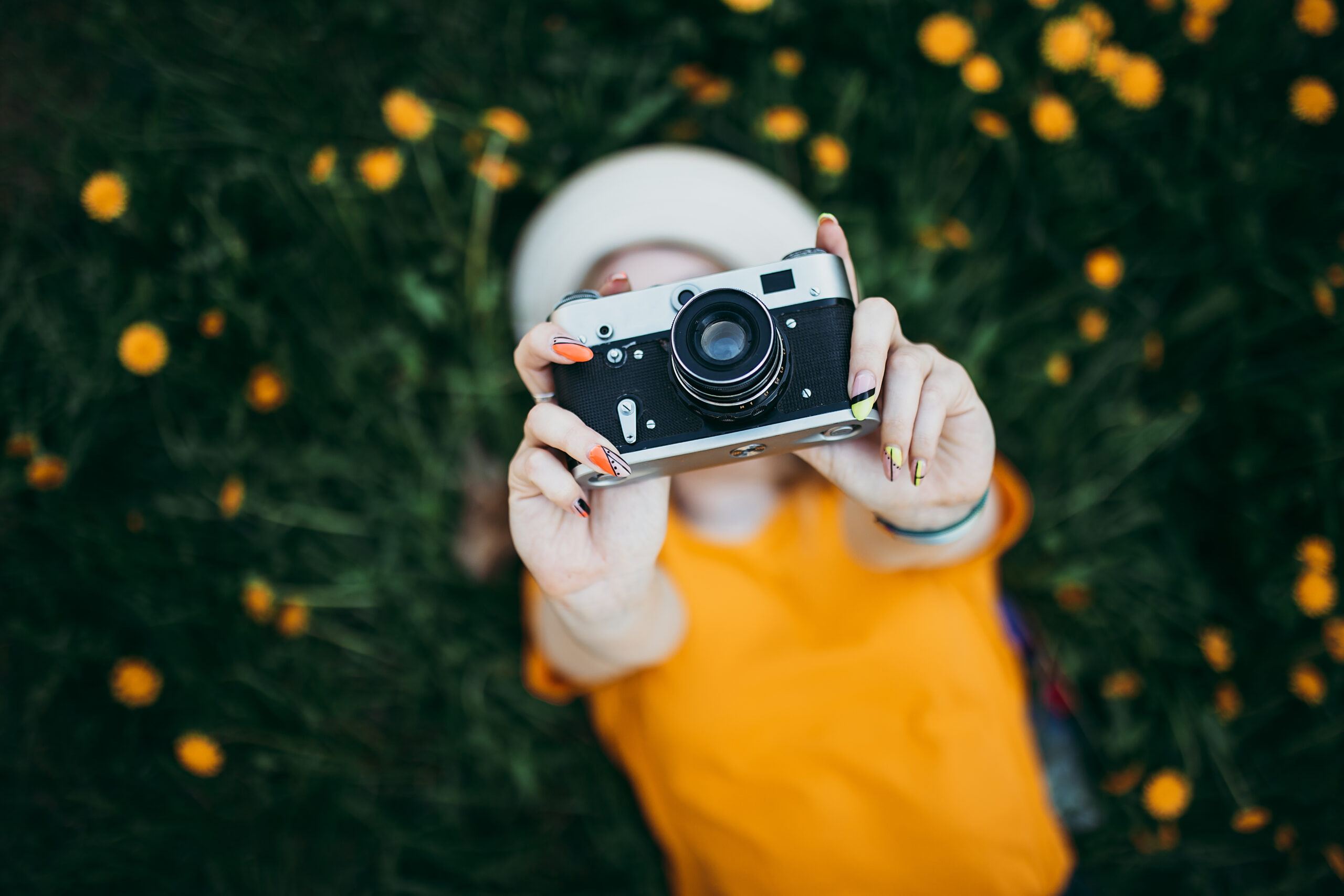 Young beautiful girl in an orange T-shirt holding retro camera in her hands lying on the lawn where dandelions grow, flowering dandelions